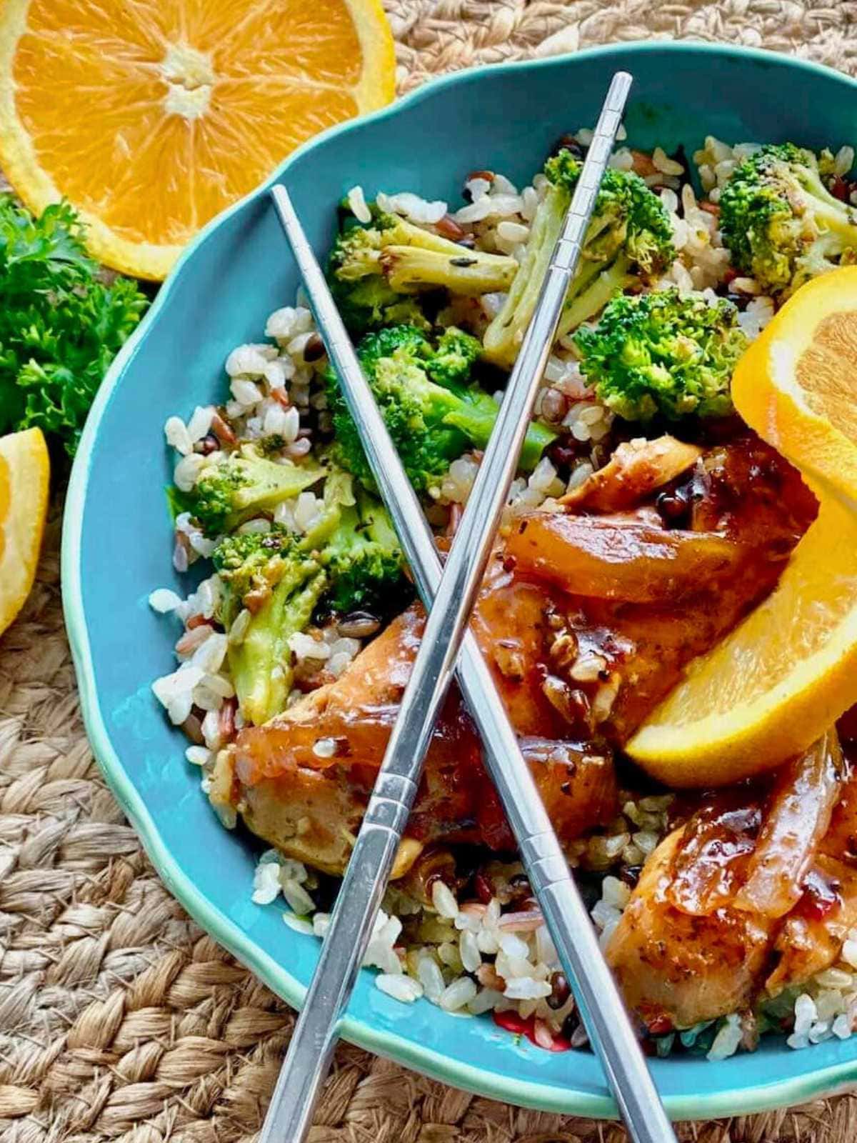 A bowl of rice with broccoli, glazed chicken, orange slices, and metal chopsticks, on a woven placemat.