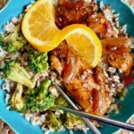 A blue plate with rice, broccoli, glazed chicken, and two orange slices, with chopsticks on the side.