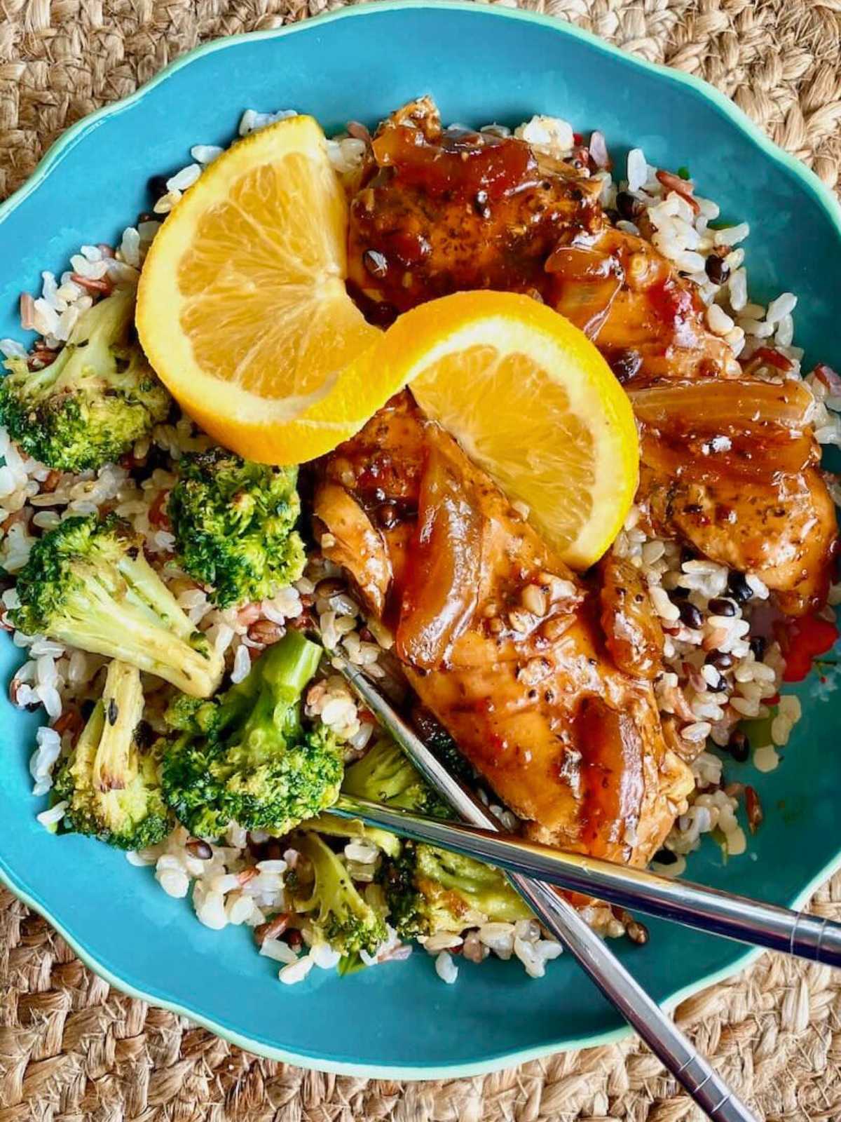 A blue plate with rice, broccoli, glazed chicken, and two orange slices, with chopsticks on the side.
