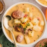 A bowl of creamy sausage and potato soup, topped with shredded cheese and herbs, with a spoon beside it.