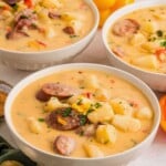 Three bowls of creamy sausage and potato soup garnished with herbs and shredded cheese on a light surface.