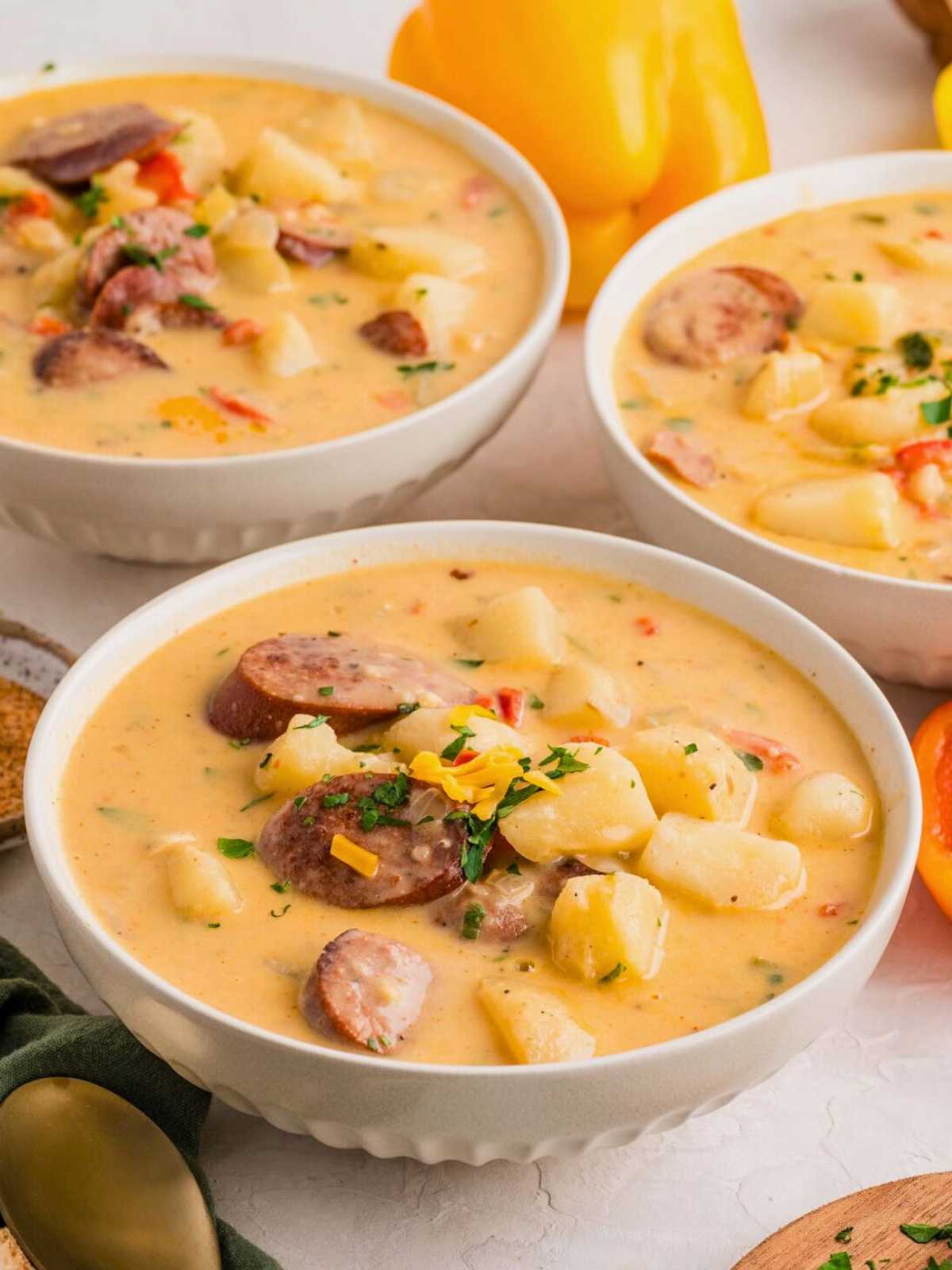 Three bowls of creamy sausage and potato soup garnished with herbs and shredded cheese on a light surface.
