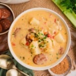 A bowl of creamy sausage and potato soup, garnished with herbs, next to fresh celery and gold spoons.
