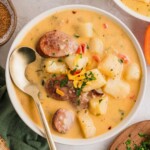 Creamy potato soup with sausage slices, cheese, and herbs in a white bowl with a gold spoon.
