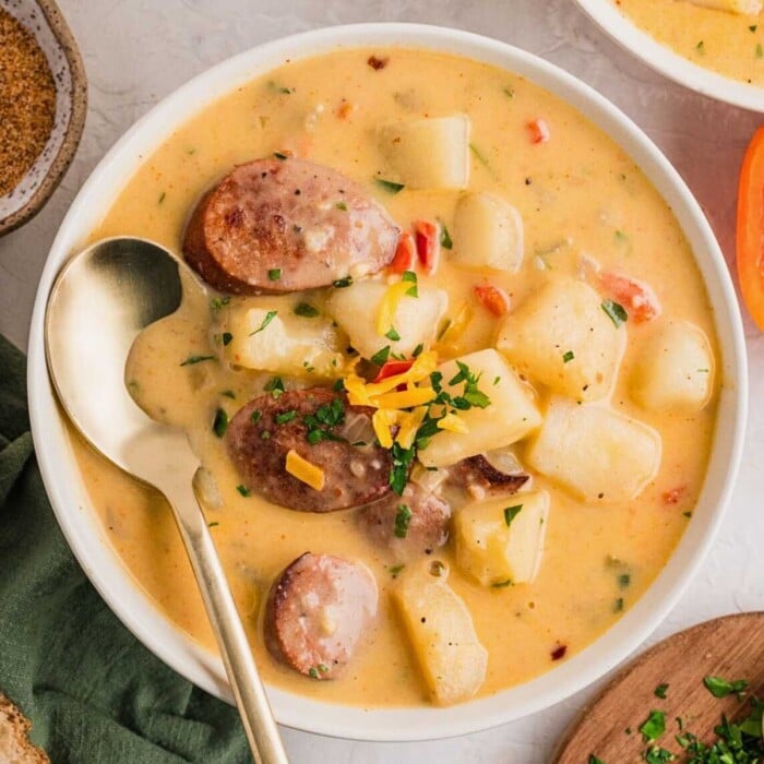 Creamy potato soup with sausage slices, cheese, and herbs in a white bowl with a gold spoon.