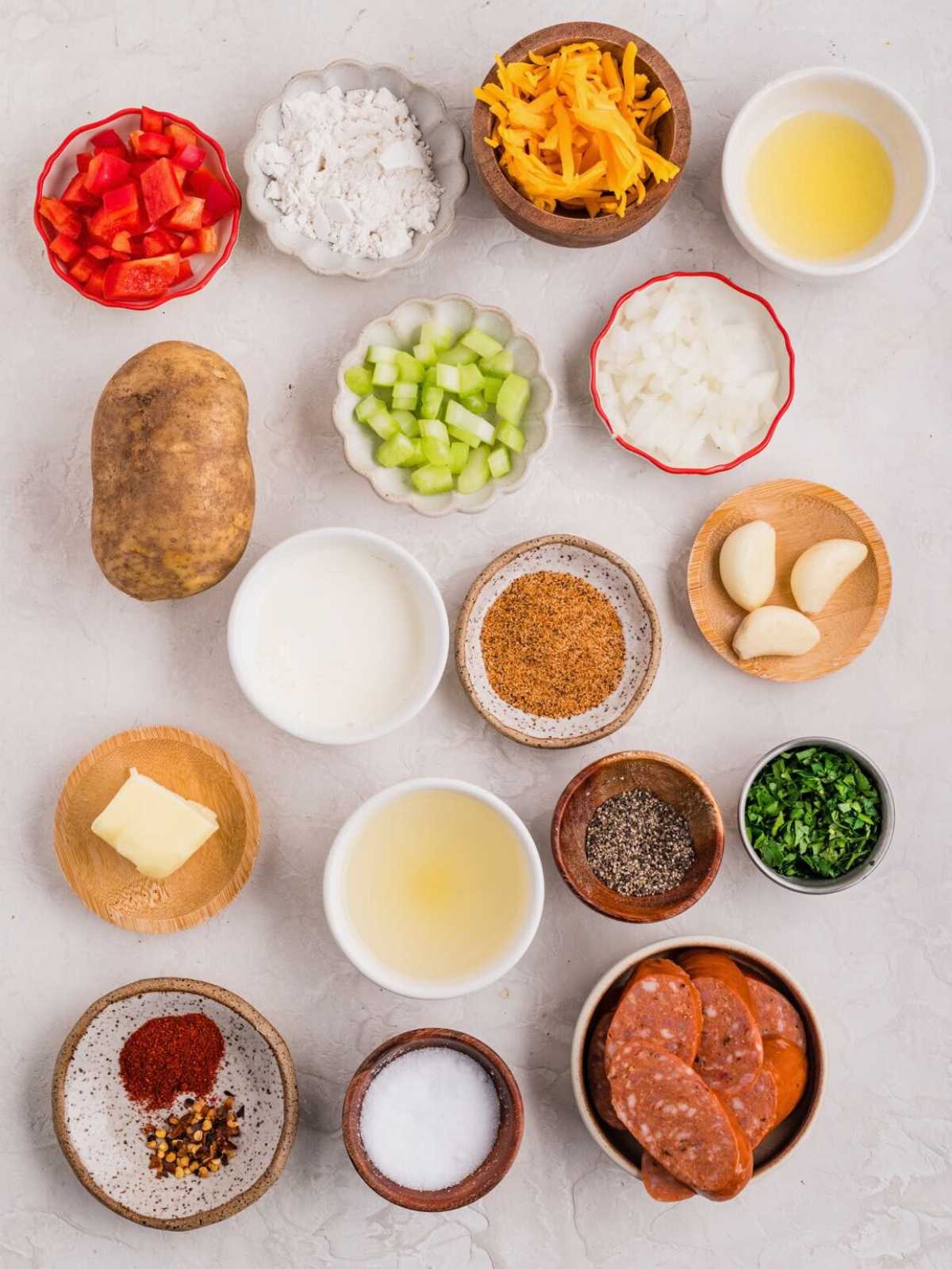 Assorted ingredients for soup, including potato, sausage, vegetables, spices, cheese, and liquids on a white surface.