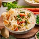 A bowl of creamy chicken and vegetable soup topped with torn biscuit pieces, surrounded by fresh vegetables.