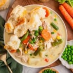Bowl of creamy chicken stew with carrots, peas, green beans, biscuits, and fresh vegetables around it.