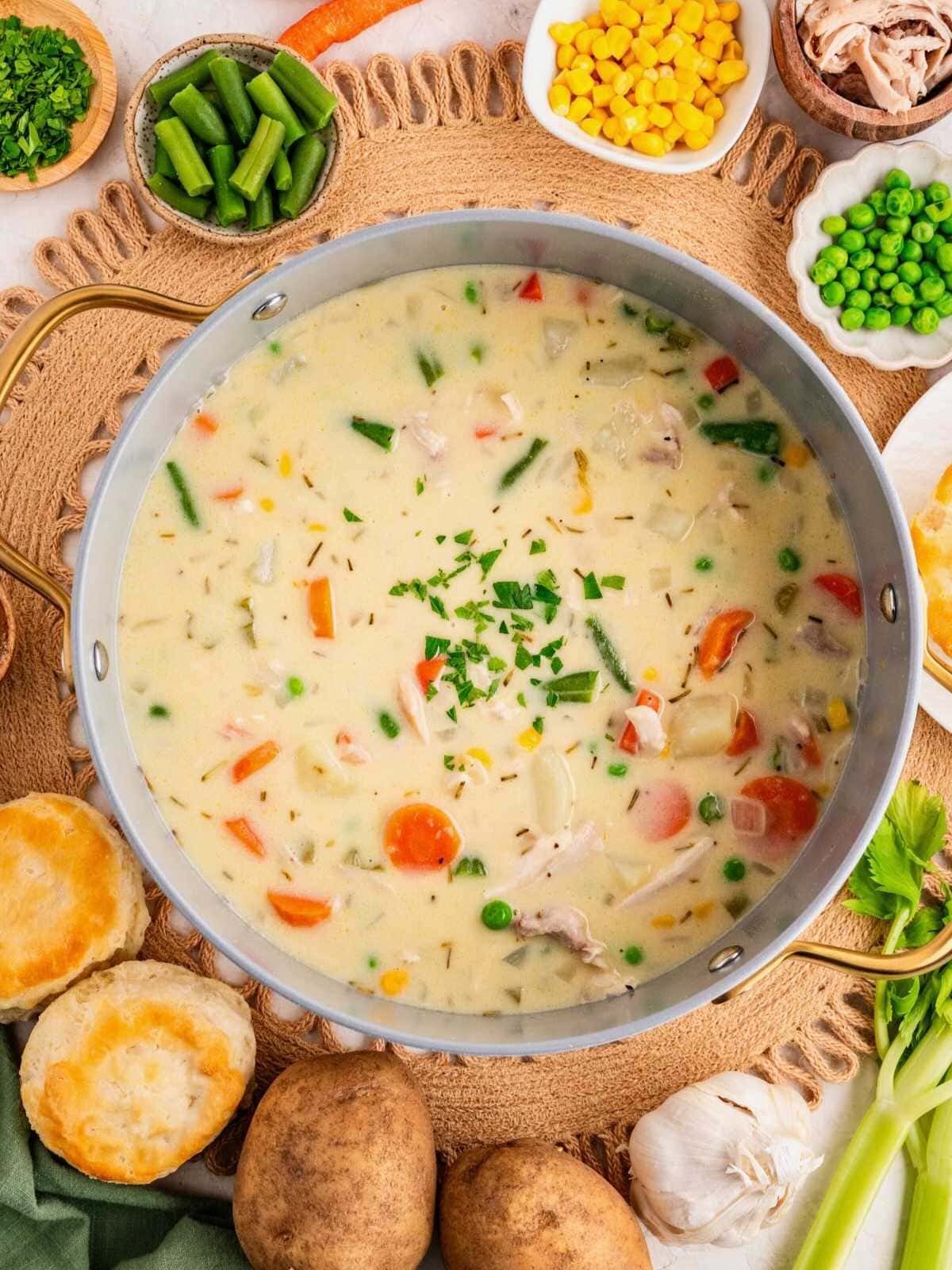 A pot of creamy chicken soup with vegetables, surrounded by biscuits, potatoes, and fresh ingredients.