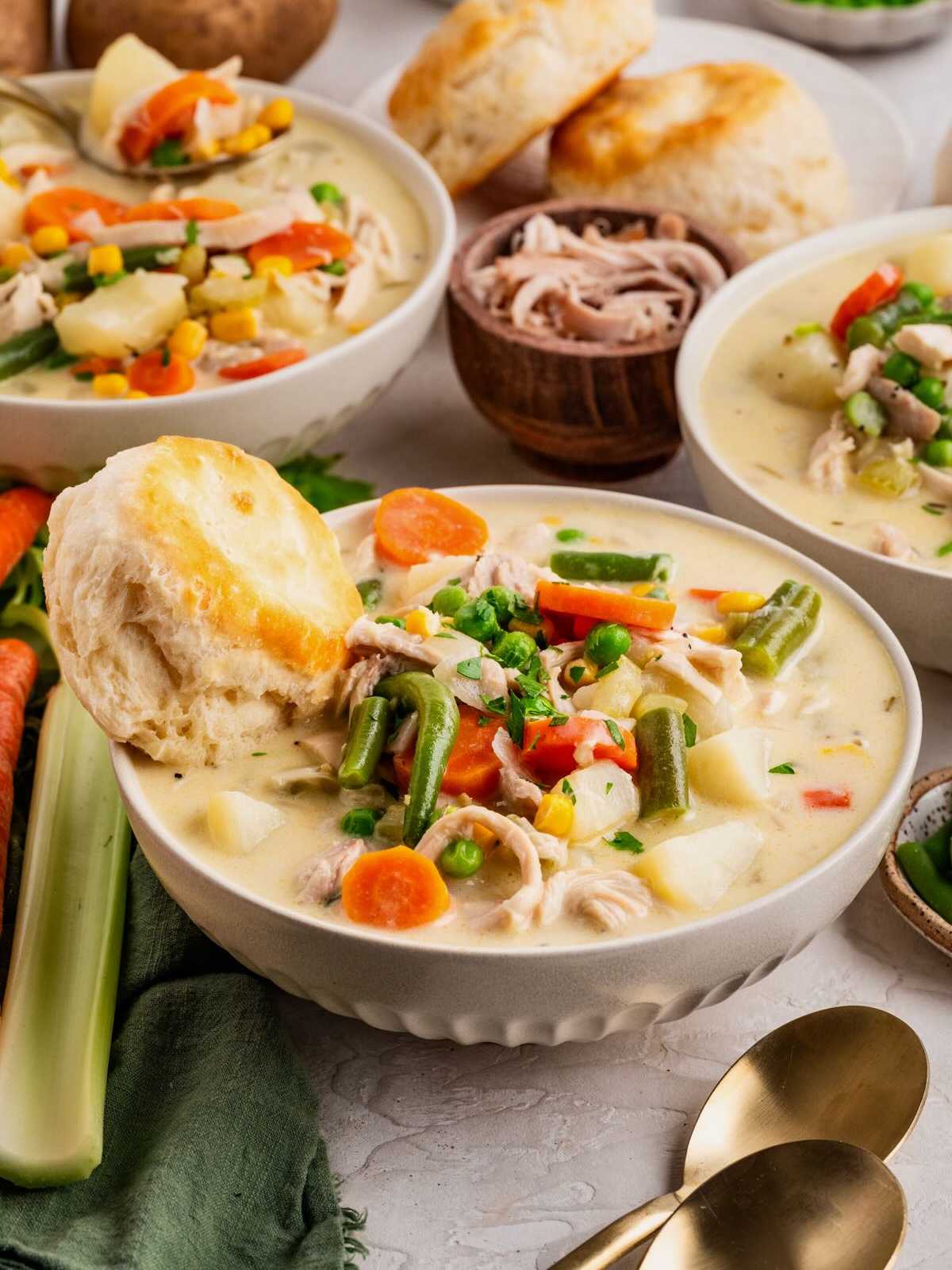 A bowl of creamy chicken and vegetable soup with a biscuit, surrounded by fresh vegetables and utensils.