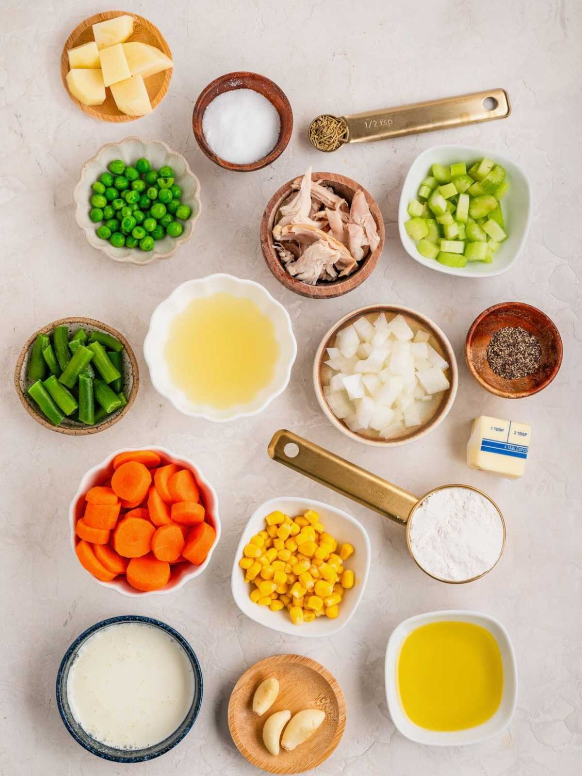 Bowls of chopped vegetables, chicken, peas, corn, seasonings, butter, flour, garlic, olive oil, and broth on a counter.