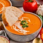 Bowl of red pepper tomato soup with a swirl of cream, garnished with herbs and a slice of bread.