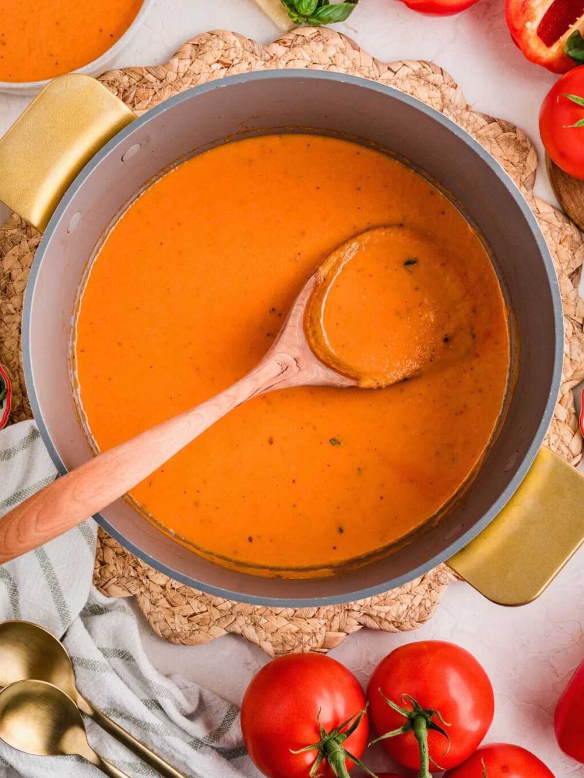 A pot of red pepper tomato soup with a wooden spoon, surrounded by fresh tomatoes and kitchen utensils.
