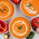 Three bowls of red pepper tomato soup with fresh basil, surrounded by tomatoes, peppers, garlic, and spoons.