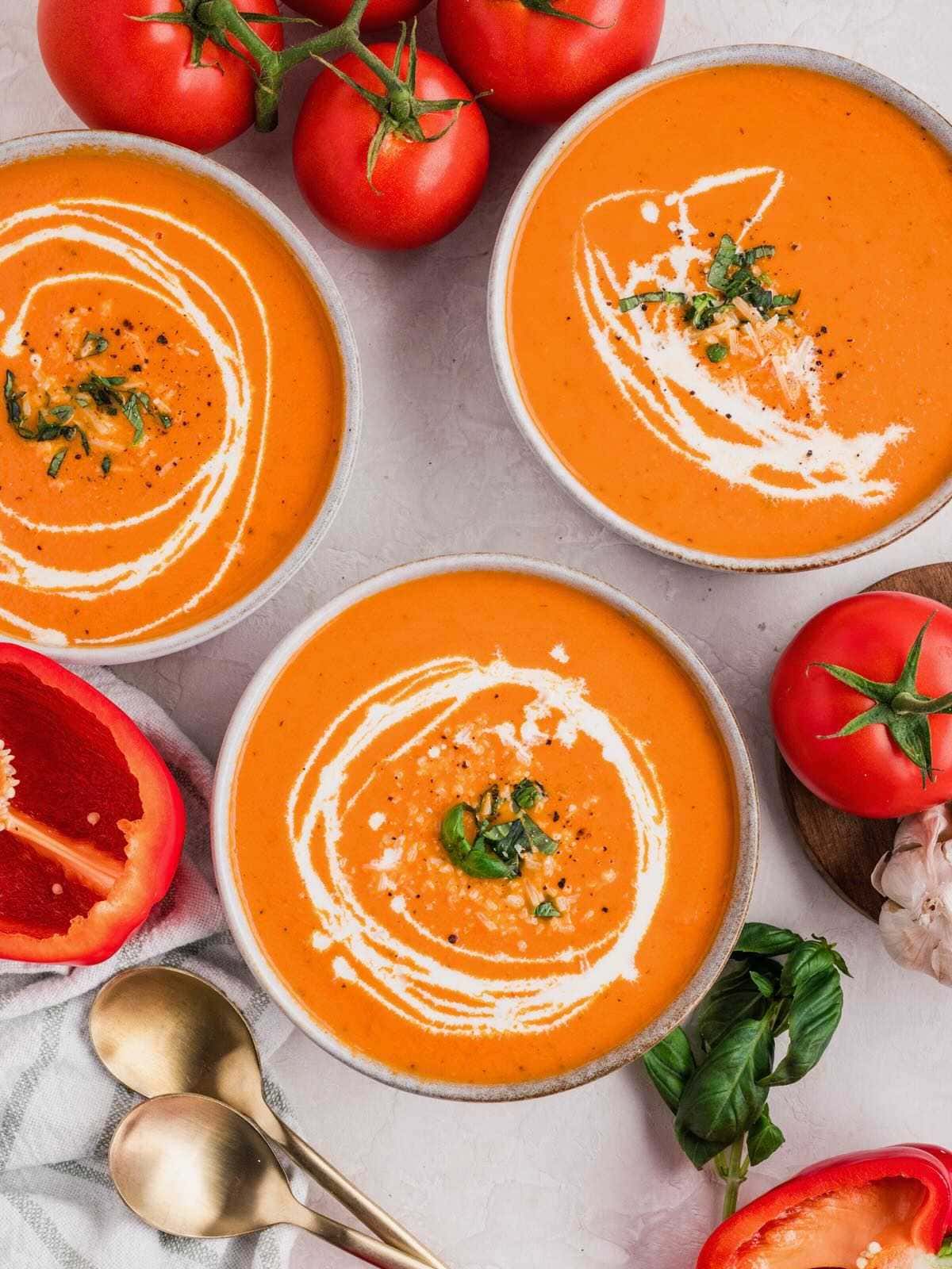 Three bowls of red pepper tomato soup with fresh basil, surrounded by tomatoes, peppers, garlic, and spoons.