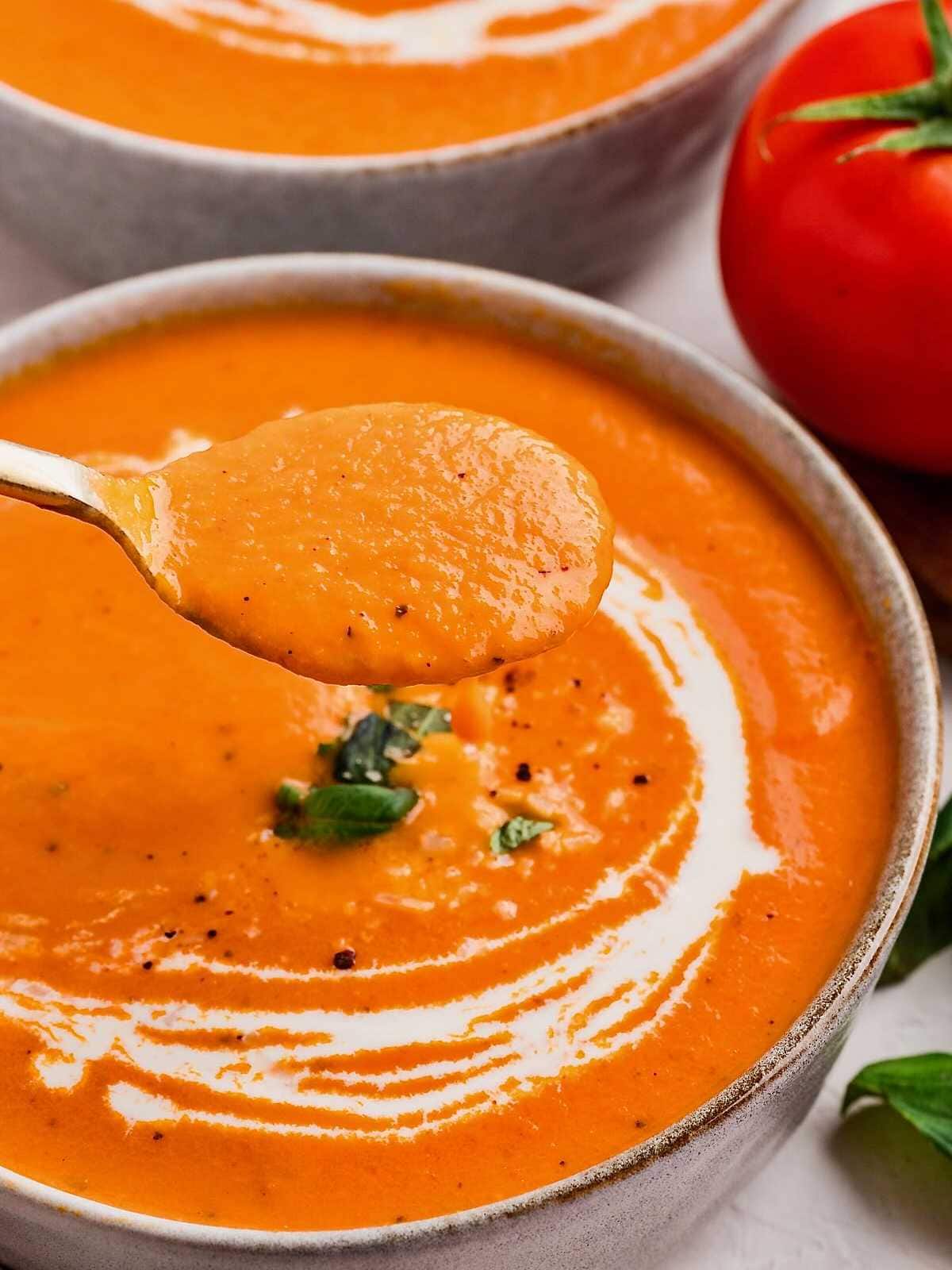 A bowl of creamy red pepper tomato soup with a swirl of cream and fresh herbs, and a spoonful held above.