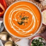 Bowl of creamy tomato soup topped with cream and herbs, with spoons and garnishes on the side.