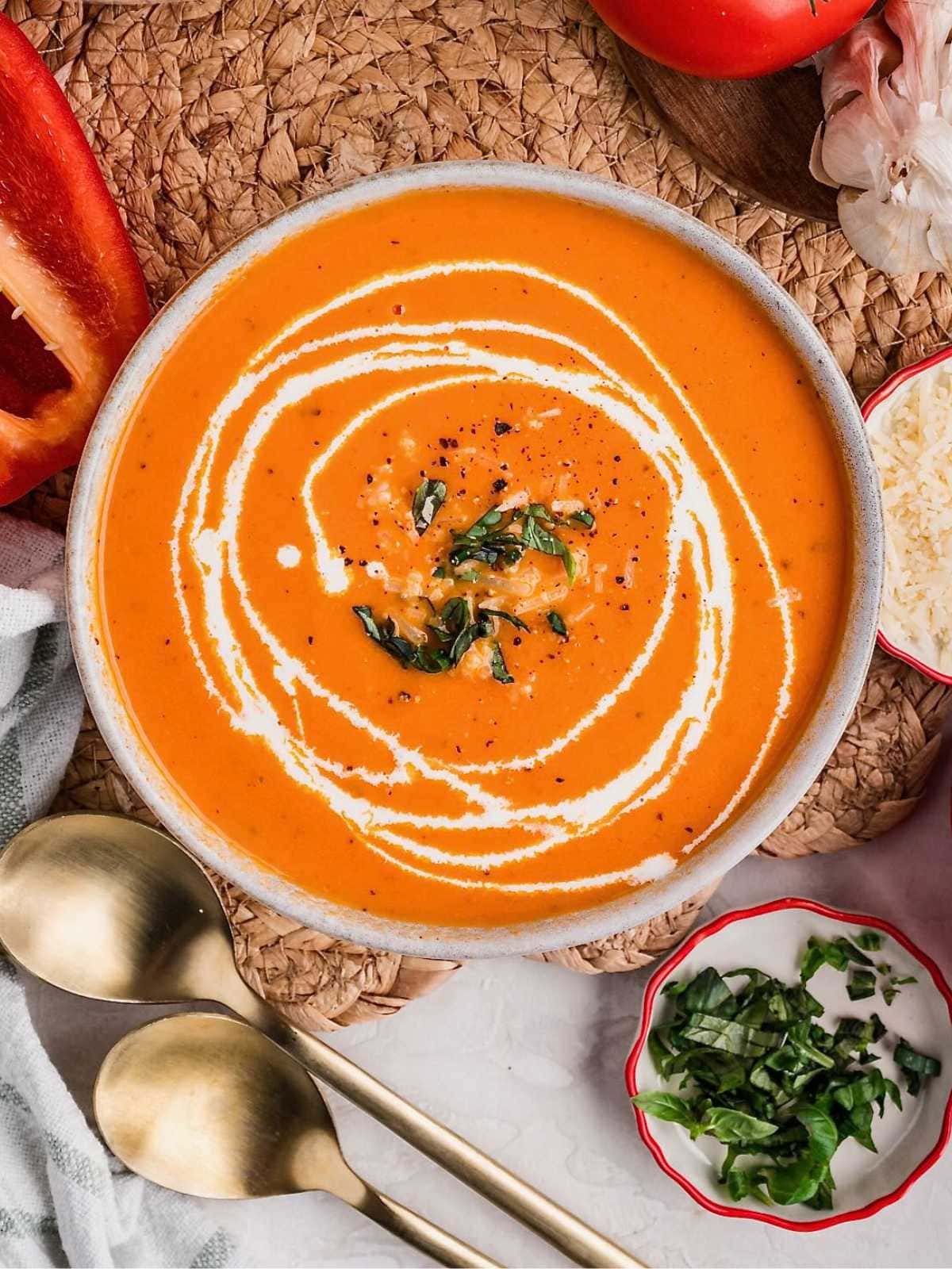 Bowl of creamy tomato soup topped with cream and herbs, with spoons and garnishes on the side.