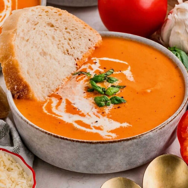 Bowl of creamy roasted red pepper tomato soup with a swirl of cream and a slice of bread, garnished with fresh basil.