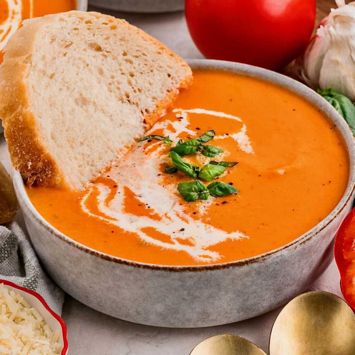 Bowl of creamy roasted red pepper tomato soup with a swirl of cream and a slice of bread, garnished with fresh basil.