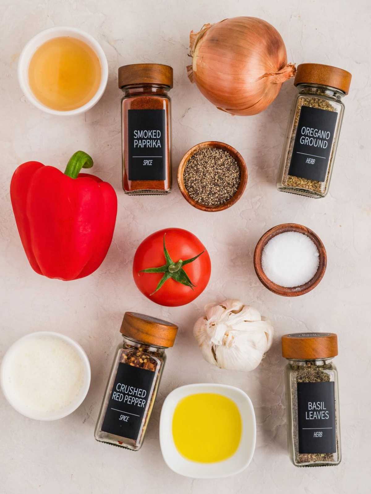 Assorted spices, vegetables, and seasonings arranged on a light surface, including onion, pepper, and tomato.