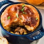 Beef stew pot pie with golden puff pastry crust in a blue ramekin, garnished with chopped herbs, next to bread slices.