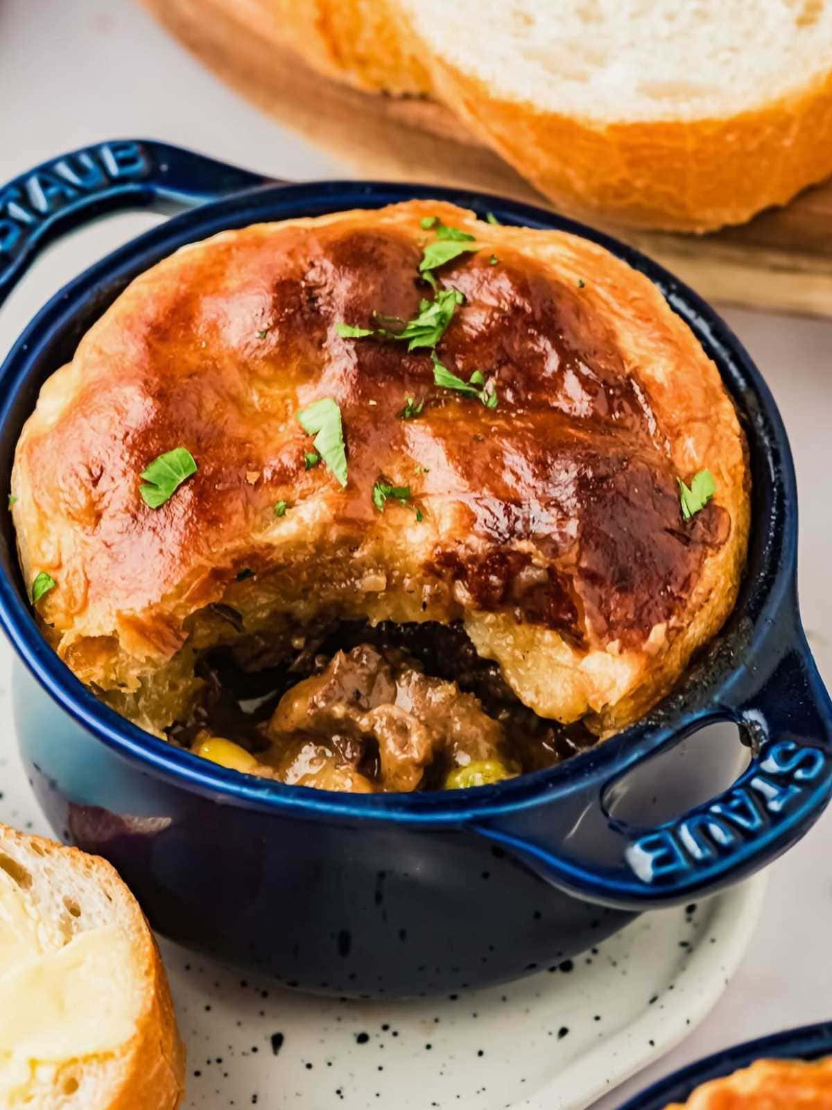 Beef stew pot pie with golden puff pastry crust in a blue ramekin, garnished with chopped herbs, next to bread slices.