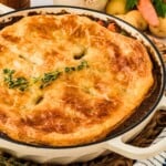 Golden, flaky pot pie topped with herbs in a white dish, surrounded by fresh vegetables and herbs.