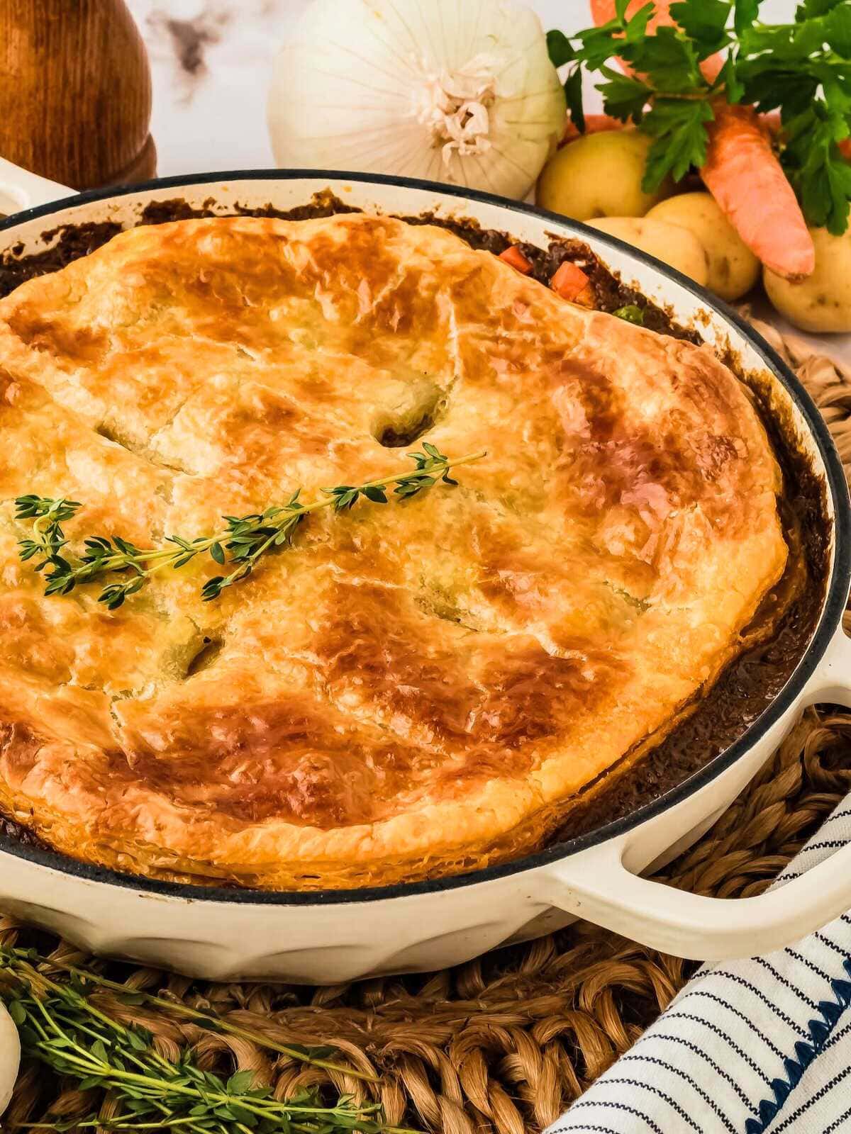 Golden, flaky pot pie topped with herbs in a white dish, surrounded by fresh vegetables and herbs.