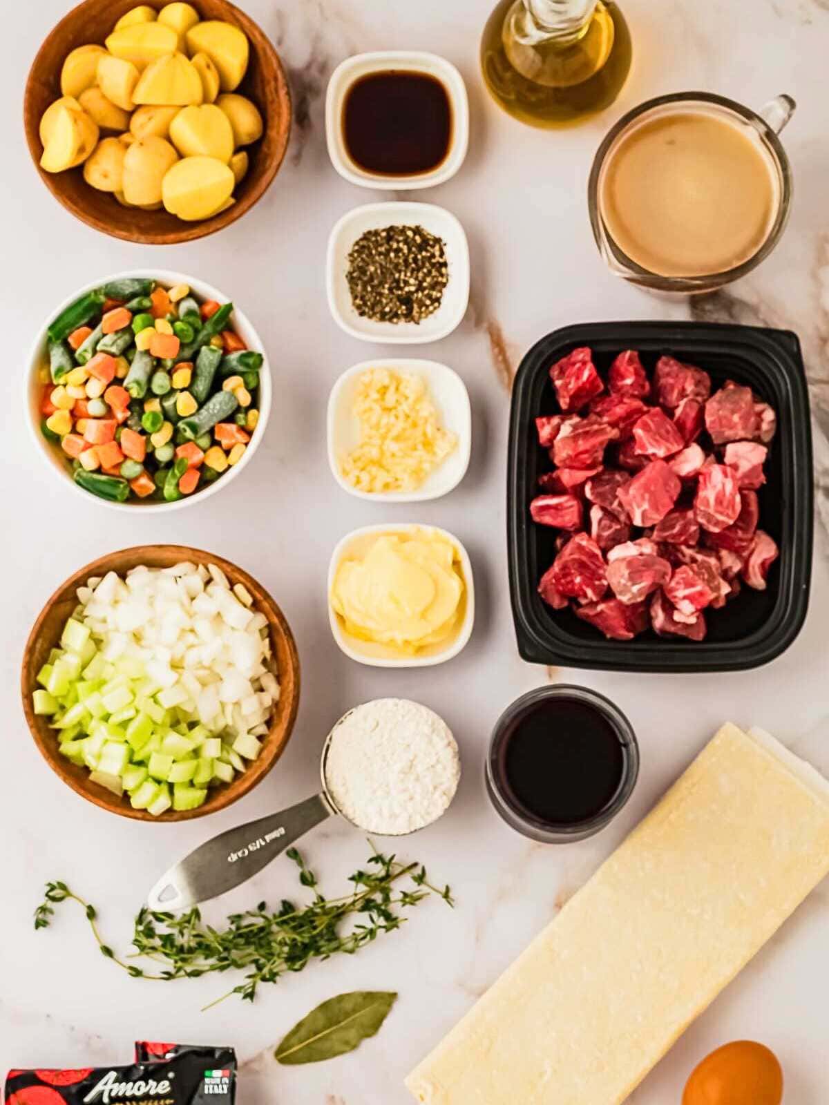 Ingredients featuring beef, vegetables, potatoes, broth, seasonings, butter, wine, flour, puff pastry, and oil on a counter.