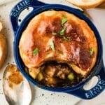 Individual beef pot pie with golden pastry crust in a blue dish, garnished with herbs, beside a spoon and bread.
