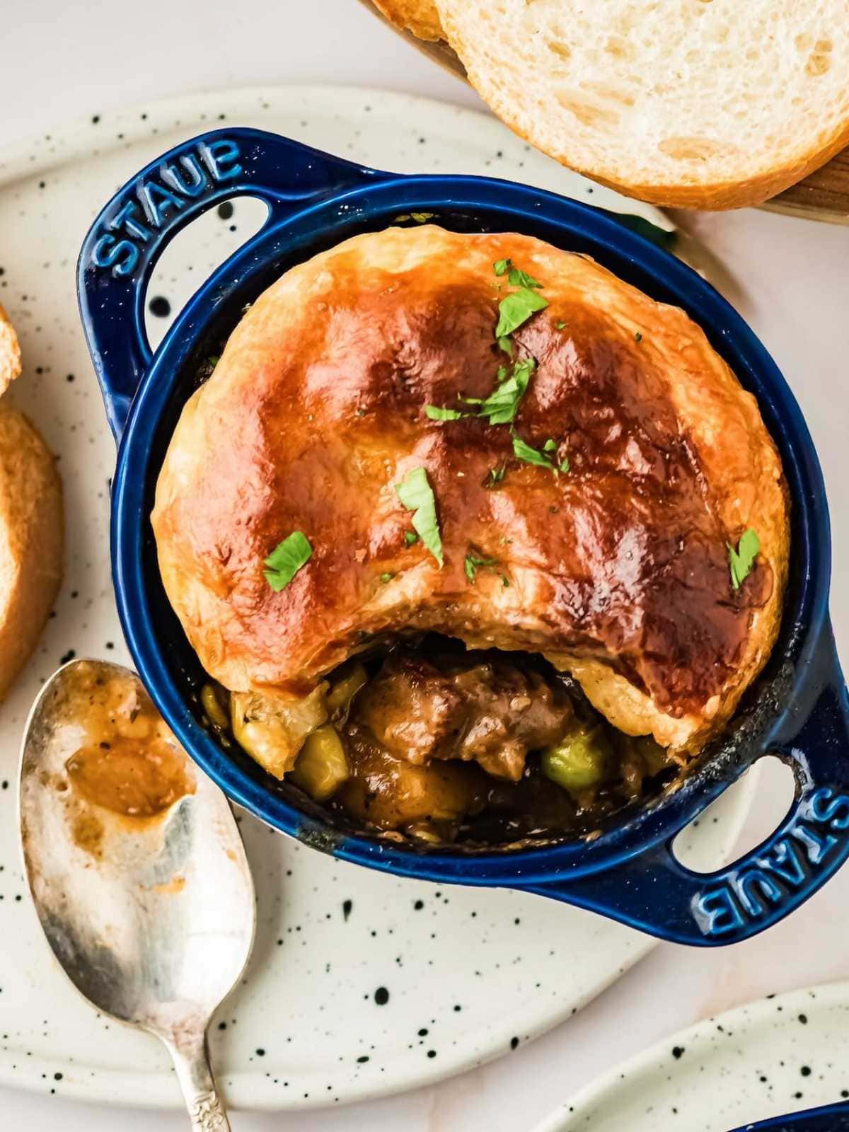 Individual beef pot pie with golden pastry crust in a blue dish, garnished with herbs, beside a spoon and bread.