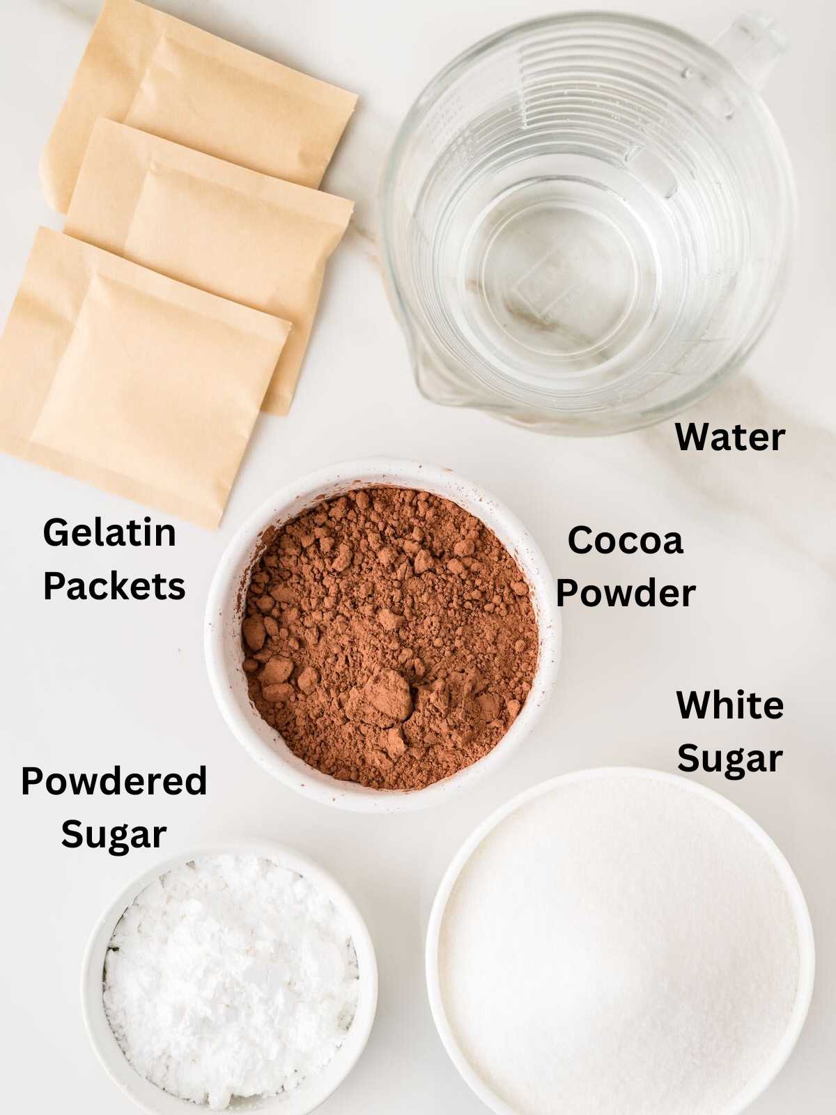 Top view of gelatin packets, cocoa powder, powdered sugar, white sugar, and a measuring cup of water on a white surface.