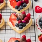 Heart-shaped cookie topped with cream, strawberries, blueberries, raspberries, chocolates, and tiny pink flowers.