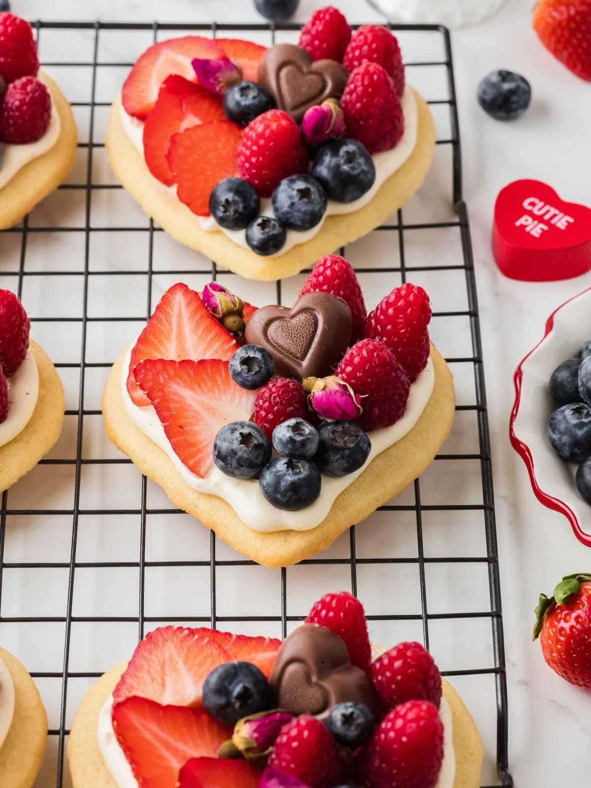 Heart-shaped cookie topped with cream, strawberries, blueberries, raspberries, chocolates, and tiny pink flowers.