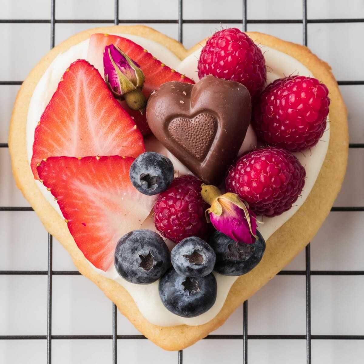 Heart-shaped cookie with cream, strawberries, raspberries, blueberries, a chocolate heart, and rosebuds on top.