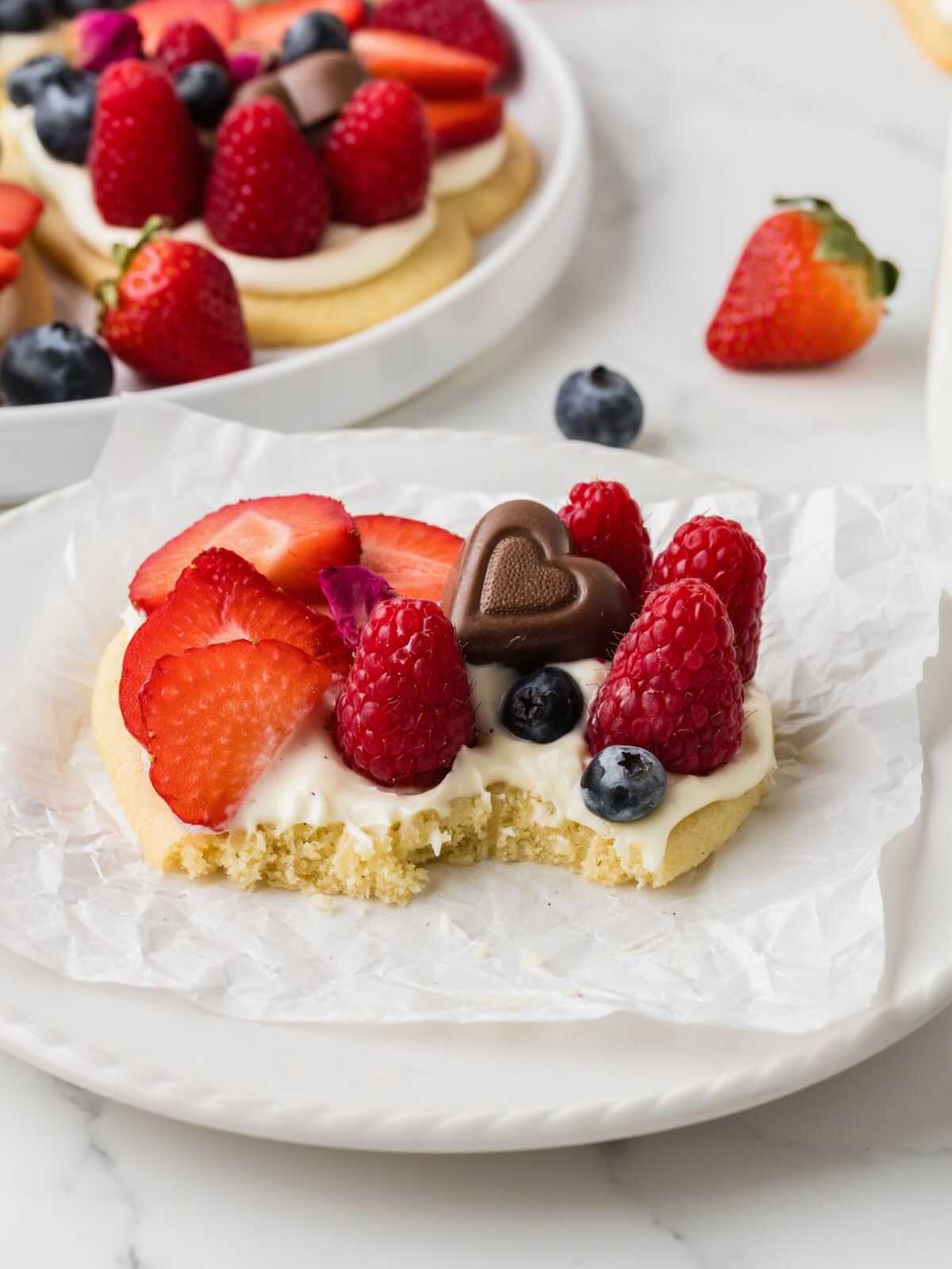 A fruit-topped sugar cookie with a bite taken out, decorated with berries and a chocolate heart.