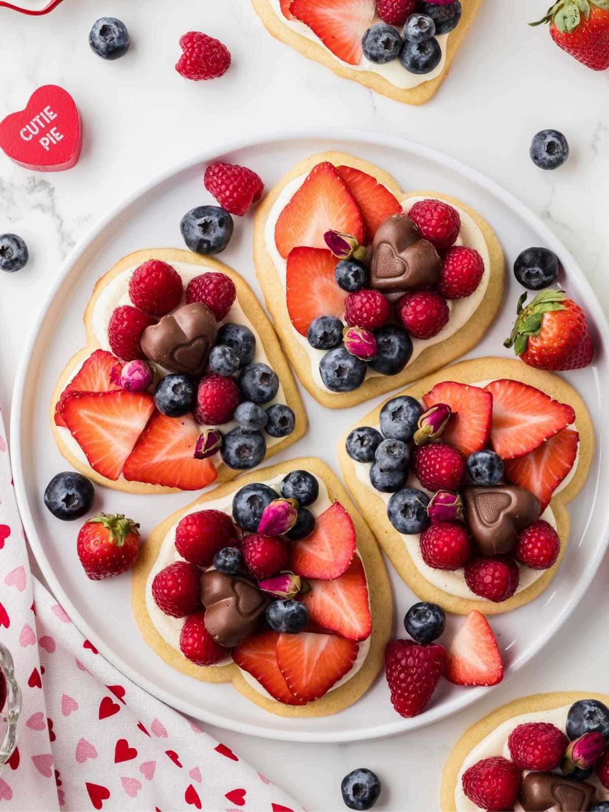 Heart-shaped cookies topped with strawberries, blueberries, raspberries, and chocolate, arranged on a white plate.