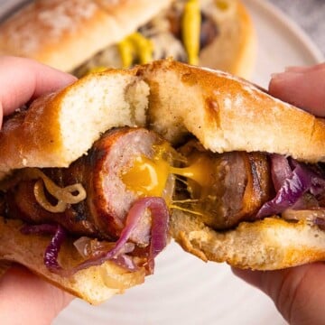 Hands holding a sausage sandwich with melted cheese, caramelized onions, and a toasted bun.