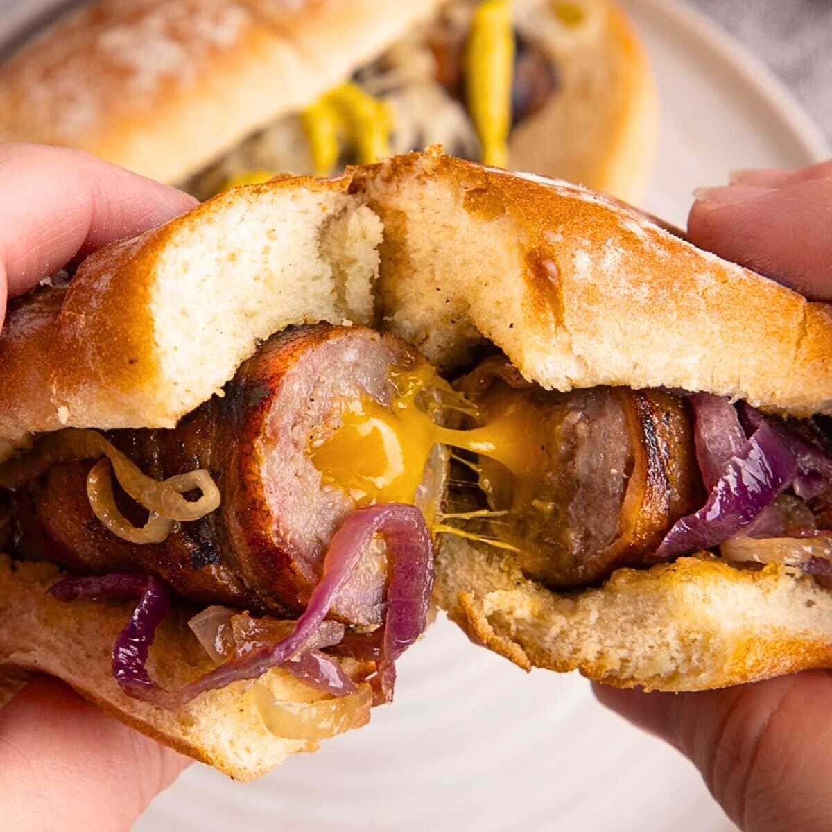 Hands holding a sausage sandwich with melted cheese, caramelized onions, and a toasted bun.