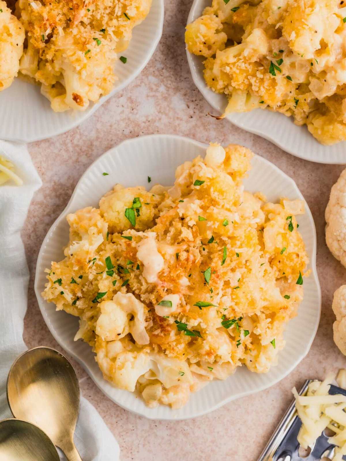 Plates of baked cauliflower cheese topped with breadcrumbs, garnished with chopped herbs, on a light surface.