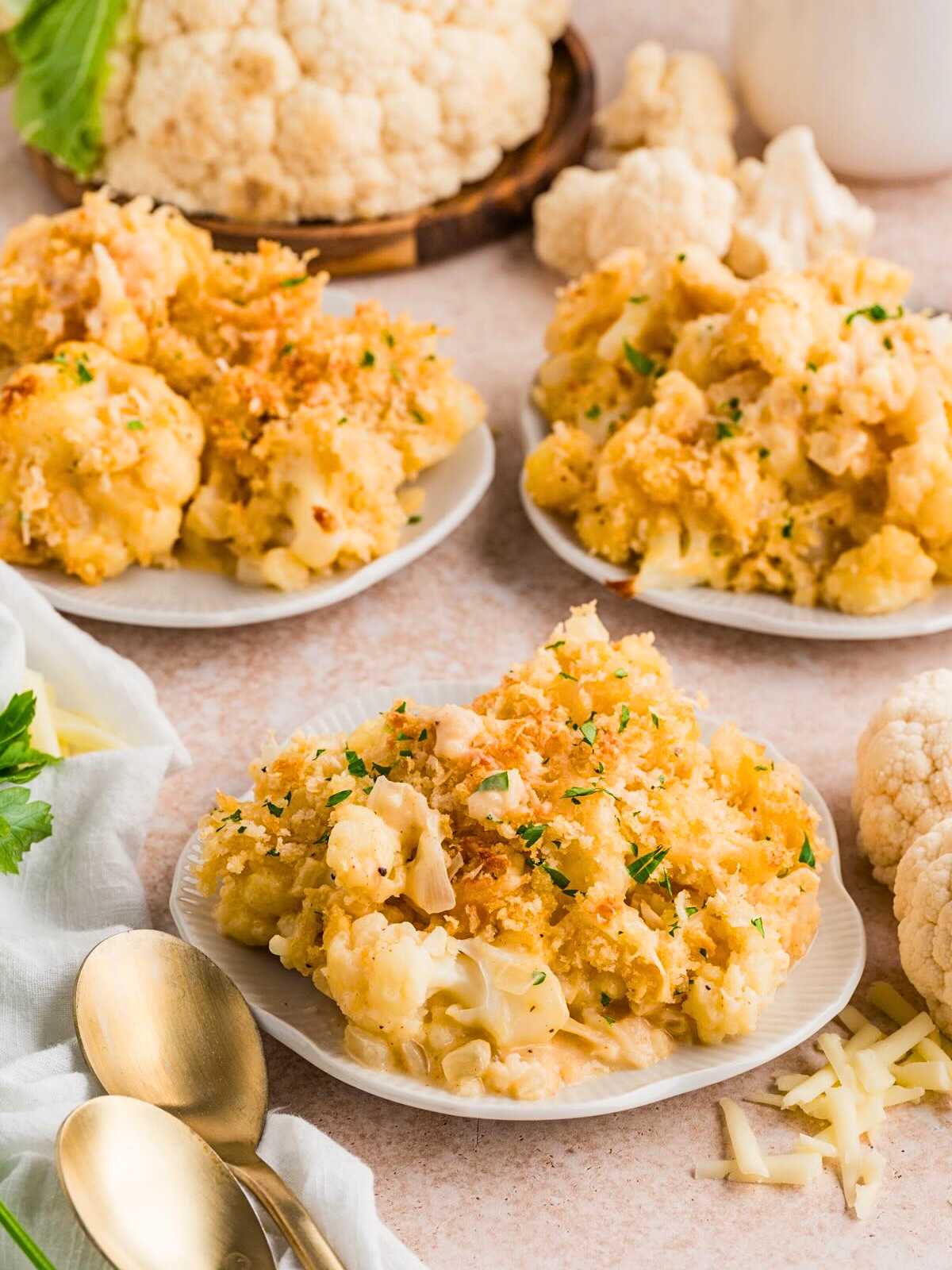 Plates of baked cauliflower gratin with golden breadcrumbs, surrounded by fresh cauliflower and herbs.