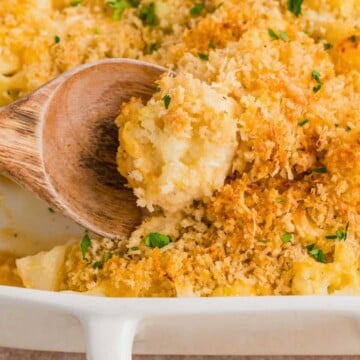 A wooden spoon scoops cheesy cauliflower casserole with crispy breadcrumb topping from a white baking dish.