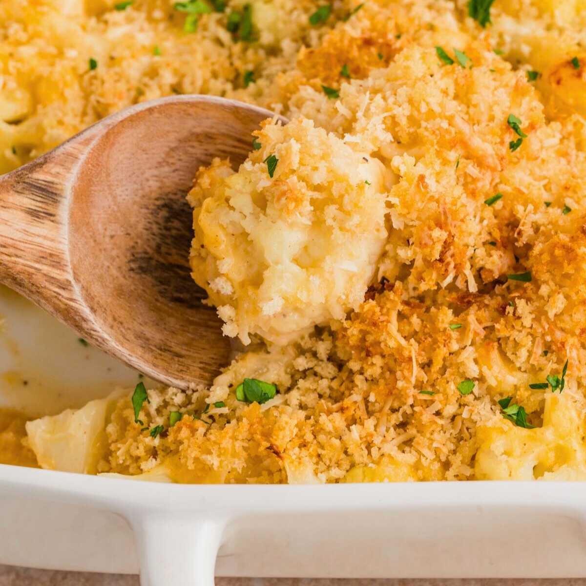 A wooden spoon scoops cheesy cauliflower casserole with crispy breadcrumb topping from a white baking dish.