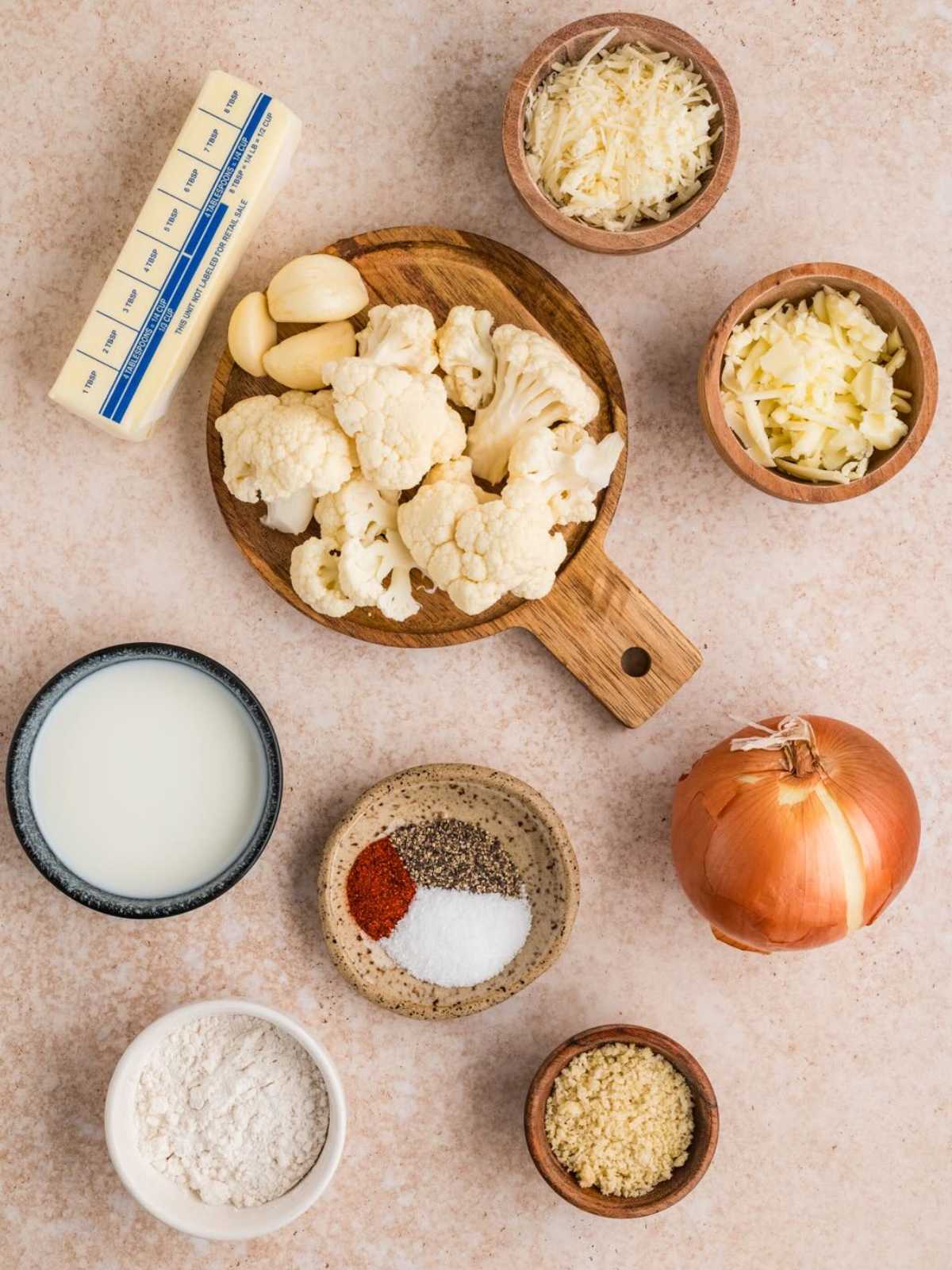 Cauliflower, butter, garlic, cheese, onion, milk, flour, breadcrumbs, and spices arranged on a light surface.
