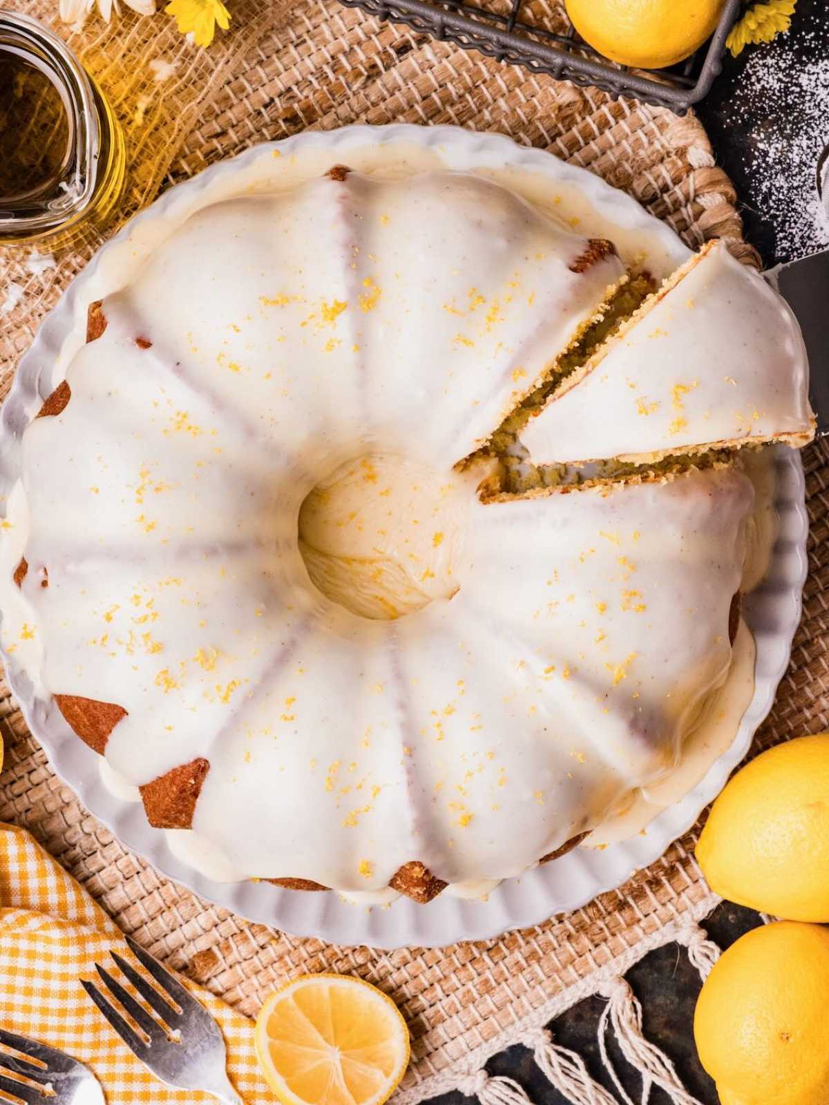 Bundt cake with white icing and lemon zest, one slice cut, surrounded by lemons and utensils.