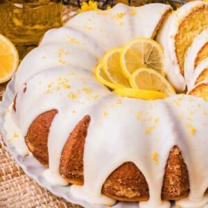 Bundt lemon cake with white icing, lemon slices, and zest on top, served on a white plate.