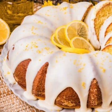 Bundt lemon cake with white icing, lemon slices, and zest on top, served on a white plate.