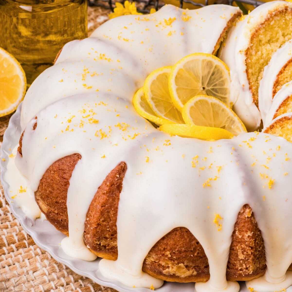 Bundt lemon cake with white icing, lemon slices, and zest on top, served on a white plate.