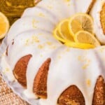 A lemon bundt cake with white icing, topped with lemon slices and zest, on a white plate.